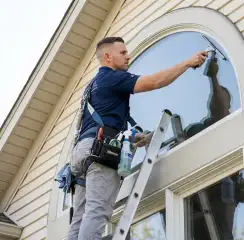 Professional window cleaner on a ladder using a squeegee to clean an exterior arched window, Houston, TX.